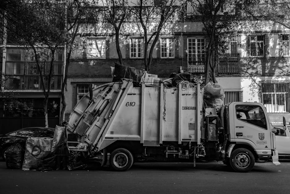 A black and white image of a waste collection truck parked on a residential street in Wennington, with trees and multi-storey apartment buildings in the background. The truck, associated with the local council, is loaded with various bulky household waste items, including large cardboard boxes, plastic bags filled with rubbish, and wooden furniture parts, some wrapped in plastic or fabric coverings. The vehicle is positioned parallel to the curb, with its rear cargo area open and filled with discarded items prepared for removal, reflecting a home clearance or furniture transport process. The street surface is visible beneath the truck, and the lighting indicates daytime, with shadows cast by the trees and nearby structures. The company's focus on removals and waste collection is subtly implied through the scene of loading and waste disposal activities, relevant for residents managing bulky waste as per Wennington council rules.