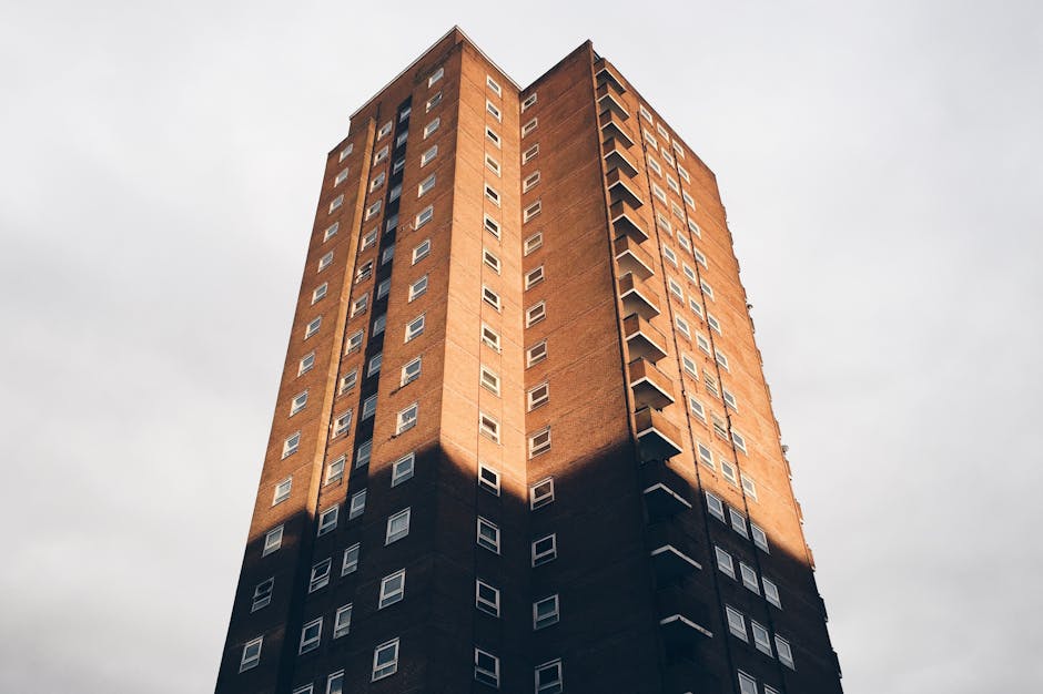 Photograph of a tall, multi-storey brick residential building with numerous small, rectangular windows arranged in a grid pattern. The building has a prominent vertical line of balconies on one side, each with a small railing, extending from the lower to the upper floors. The lower portion of the building casts a shadow on the middle sections, creating a contrast between light and dark areas. The building is situated against a cloudy sky, and the perspective emphasizes its height and verticality. This image, associated with house removals and moving services, illustrates an urban dwelling where home relocation and furniture transport might take place, with the structure interior or exterior ready for a professional moving process by companies like Man with Van Wennington.