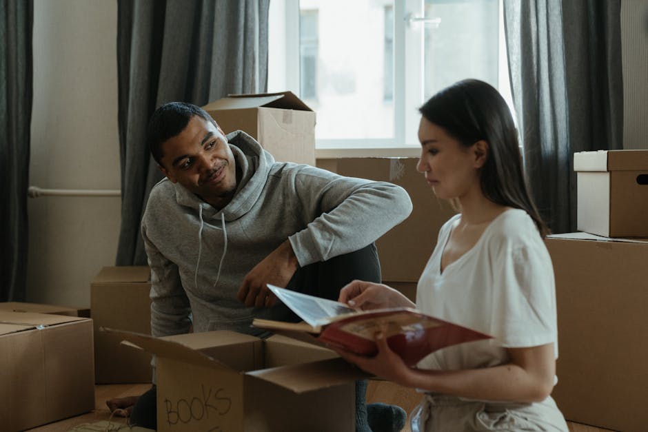 A man and a woman in a residential interior surrounded by packed cardboard boxes, some labeled with items like 'books'. The man, wearing a grey hoodie, is sitting on the floor next to a cardboard box, looking at the woman with a slight smile. The woman, dressed in a white t-shirt, is holding open a folder or document, appearing to review or read information. Natural light enters through a window behind them, illuminating the room and the ongoing home relocation process, with additional boxes stacked nearby. The setting suggests the process of packing and preparing for a move, with the individuals engaged in discussing or organizing their belongings. Man with Van Wennington, a professional removals service, may assist with such moving and furniture transport tasks, ensuring a smooth transition during house relocations, including compliance with leaseholder move rules for Wennington flats.