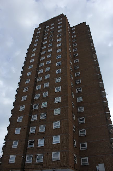 Photograph of a tall, multi-storey brick residential building with numerous small, rectangular windows arranged in a grid pattern. The building has a prominent vertical line of balconies on one side, each with a small railing, extending from the lower to the upper floors. The lower portion of the building casts a shadow on the middle sections, creating a contrast between light and dark areas. The building is situated against a cloudy sky, and the perspective emphasizes its height and verticality. This image, associated with house removals and moving services, illustrates an urban dwelling where home relocation and furniture transport might take place, with the structure interior or exterior ready for a professional moving process by companies like Man with Van Wennington.