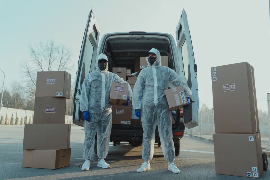 A man wearing a white t-shirt and jeans is standing on the pavement next to a white moving van with its rear doors open, engaged in a home relocation. He is in the process of unpacking or organizing c