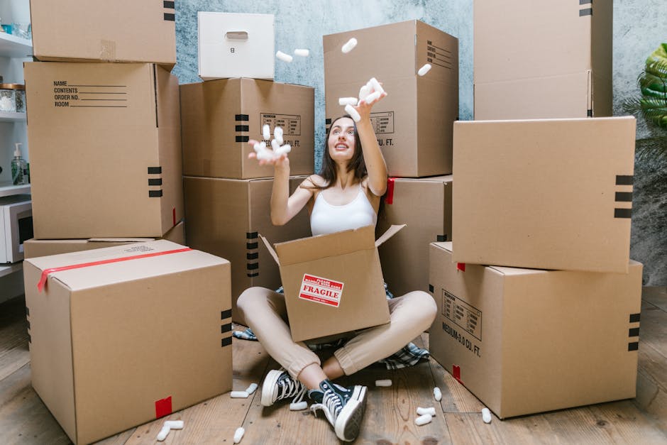 A woman sitting cross-legged on a wooden floor surrounded by numerous large cardboard boxes in an indoor setting, with some boxes sealed with red tape and others labeled with handling instructions. She is wearing a white tank top, beige pants, and striped sneakers, and is in the process of throwing packing foam peanuts into the air, smiling as part of a packing process associated with home relocations. Behind her, the room features a textured wall and a small shelf with items, indicating an active packing environment. The boxes are stacked at various heights, some to her sides and others behind her, suggesting a house removal or moving preparation. The scene is well-lit, emphasizing the organized yet busy atmosphere typical of furniture transport and packing during moves, aligned with the services offered by Man with Van Wennington.
