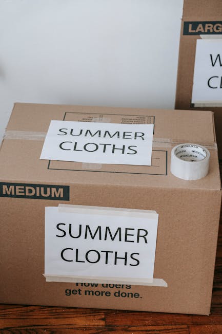 A large brown cardboard box labeled 'SUMMER CLOTHS' with a white label on the top and a smaller label on the front, situated on a wooden floor inside a property. The box is sealed with clear packing tape and has an additional piece of masking tape below the front label. Adjacent to the box, a roll of white packing tape is positioned on top of another box partially visible in the background. The environment suggests an interior space prepared for home relocation, with furniture and other packing materials likely nearby as part of the packing and moving process. The scene reflects careful organization during furniture transport and loading operations as part of house removals or a move managed by Man with Van Wennington, focusing on efficient packing of belongings before loading into a moving van.