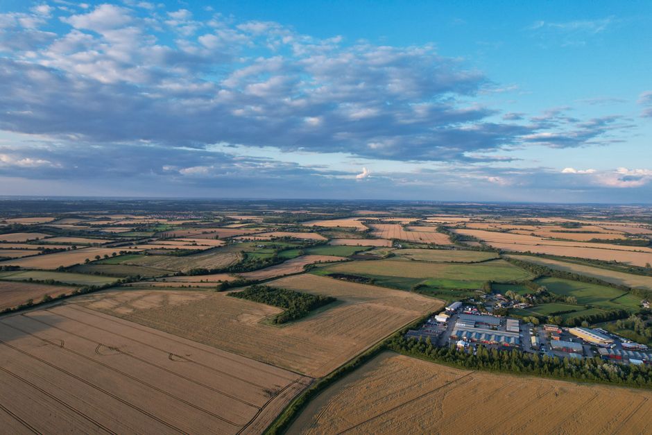 Aerial view of a rural landscape featuring large agricultural fields divided by hedgerows and narrow roads, with a commercial area consisting of multiple white and grey industrial buildings and warehouses located in the lower right section of the image. The fields display varying shades of green and brown, indicating different crops or harvest stages, and the scene is illuminated by natural daylight with a partly cloudy sky overhead. This image illustrates the expansive outdoor environment surrounding Wennington, which can be associated with house removals and the transportation of furniture or belongings during a home relocation or packing and moving process, as handled by Man with Van Wennington.