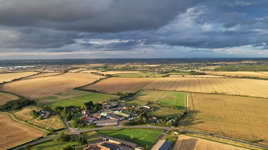 An aerial view showcasing a rural landscape near Wennington, featuring expansive fields with varying crops and grass areas, separated by hedgerows and narrow roads. In the foreground, a small cluster of residential buildings and farm structures are visible, surrounded by green lawns and trees. A narrow asphalt road curves through the scene, leading towards larger agricultural buildings and open land in the distance. The sky above is partly cloudy with dark, thick clouds casting shadows across parts of the landscape, suggesting weather changes. This image illustrates a typical countryside environment where home relocation and furniture transport services such as those provided by Man with Van Wennington may operate to assist with house removals, packing, and loading processes in rural settings.