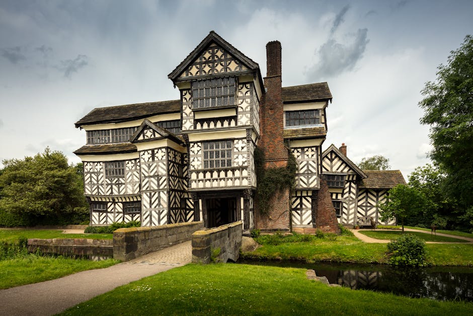 A large historic building with black-and-white timber-framed façade, multiple gabled roofs, and tall chimneys, situated behind a stone bridge and path leading to the entrance. Tall windows are visible on each floor, with some covered by wooden shutters. The surrounding area includes green lawns, mature trees, and a small pond reflecting the building, with an overcast sky overhead. This setting depicts a traditional, heritage property potentially involved in house removals or relocation services, with Man with Van Wennington prepared for furniture transport or moving logistics in the context of property transition, supported by the classic architecture and landscaped environment.