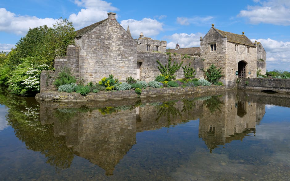 A historic stone building with a sloped, tiled roof, situated on the edge of a calm body of water that creates a clear reflection of the structure. The building features multiple gabled sections, small windows, and an arched doorway, with greenery including shrubs, flowering plants, and climbing vines along the foundation and walls. To the left, there is a large bush with white blooms, and on the right, a stone wall with an opening visible near the base. The sky above is partly cloudy with patches of blue, and the surrounding landscape appears lush and green. This scene, captured during daylight hours, presents a peaceful and picturesque setting, which is consistent with the type of location where a home relocation — possibly involving house removals or furniture transport — might take place, with the historic building serving as a notable landmark. Man with Van Wennington’s services are relevant to such relocation needs.