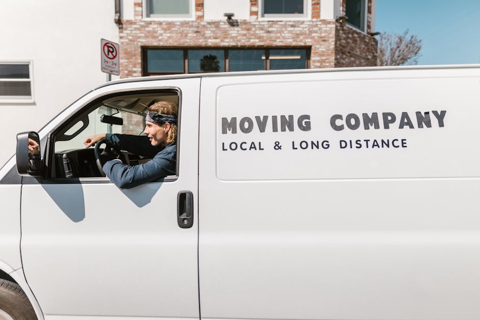 A white moving van parked on a residential street in Wennington with the company's branding, 'MOVING COMPANY LOCAL & LONG DISTANCE,' displayed on the side. Inside the vehicle, a professional mover with long hair, wearing a headband and dark jacket, is seen through the open driver's window, preparing to load furniture or boxes. The background features a brick building with large windows and a tree, along with a no-parking sign on the pavement indicating parking rules for vans and moving vehicles. The scene captures the loading process typical of home relocation services offered by Man with Van Wennington, including furniture, cardboard boxes, and packing materials visible in the vehicle or nearby, emphasizing efficient transportation logistics during the moving process.