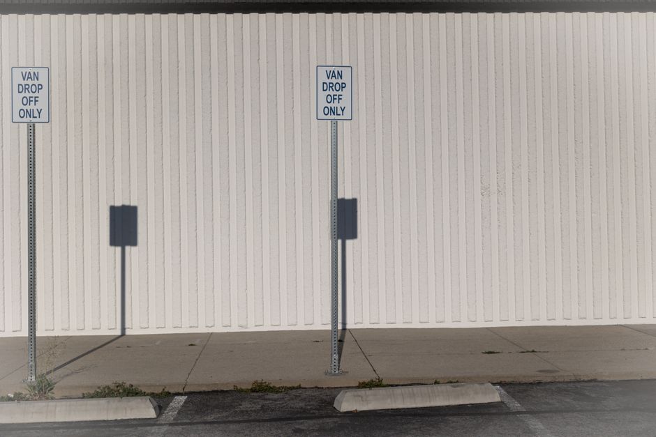 A plain white wall with vertical paneling serves as the background for two designated van drop-off and parking signs mounted on metal posts. The signs display the message 'VAN DROP OFF ONLY' in blue text on a white background. The middle sign is positioned directly in front of a space on the pavement, where a large vehicle, likely a van involved in a home relocation or furniture transport service, is parked nearby. The pavement below shows slight cracks and patches of grass growing through. Shadows cast by the signs and their posts indicate the light source is coming from the left side of the image. The environment appears clean and organized, typical of designated loading or unloading zones used for removals or moving services. Man with Van Wennington occasionally provides such loading areas as part of its removal process, ensuring compliance with local parking and permit regulations when executing house removals and furniture transportation.