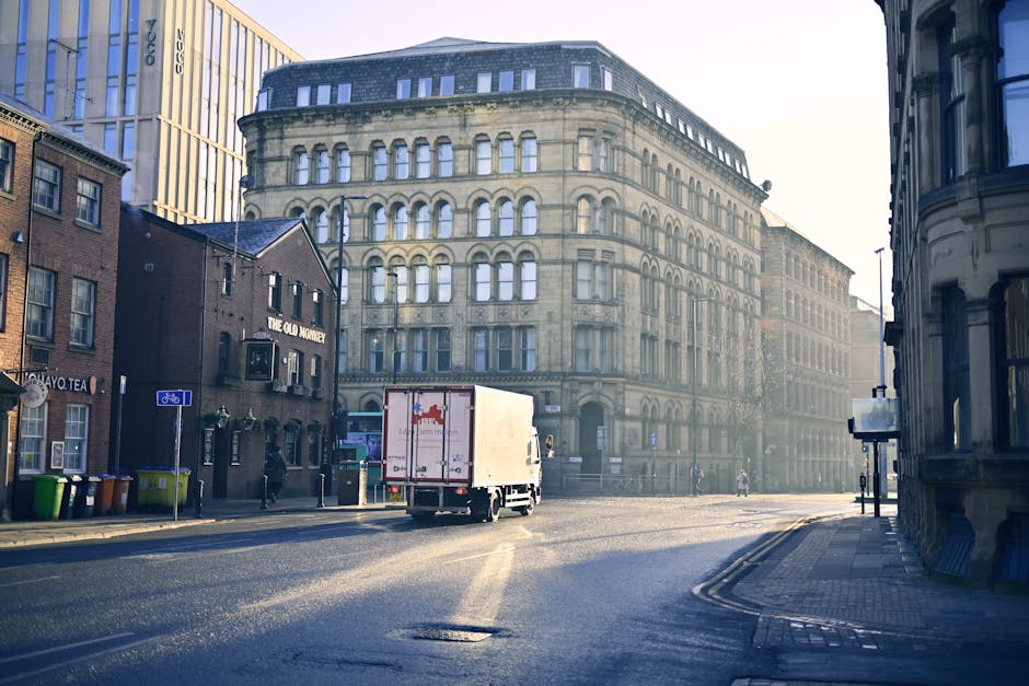 A large green box truck parked on a city street in Wennington, surrounded by tall modern office buildings and older stone structures, with a clear blue sky overhead. The truck is positioned near the c