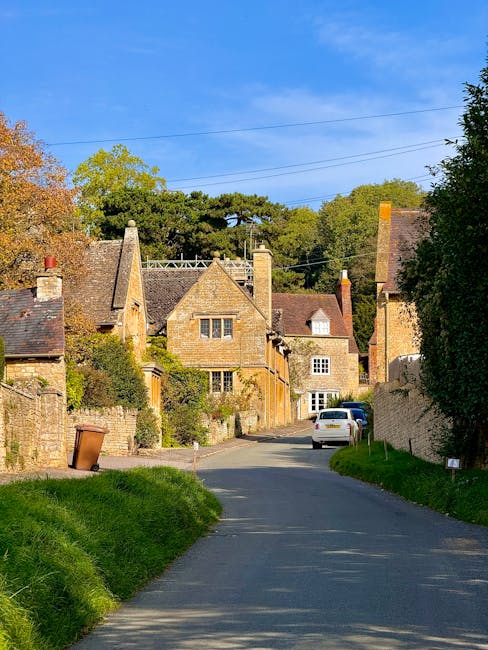 A narrow residential street in Wennington Village under a bright blue sky, with traditional stone houses featuring pitched roofs, chimneys, and small windows. The street is paved and slightly curved,